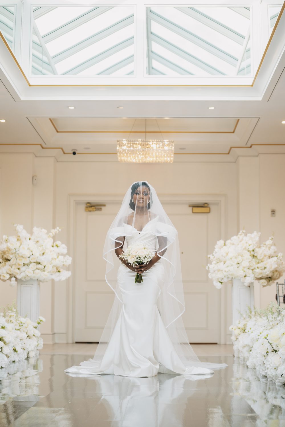 A veiled bride standing between white floral arrangements under a skylight